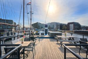 Vue d'une terrasse en bois sur un bateau amarré, avec des tables, des chaises et des vases à fleurs, entourée d'eau et de bâtiments modernes, sous un ciel lumineux où le soleil est partiellement visible. - Ekilib