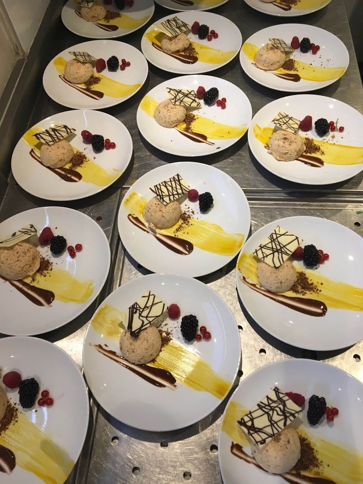 Plusieurs assiettes blanches contiennent chacune un dessert composé d'une cuillère de mousse, d'une décoration en chocolat, de filets de sauce jaune et marron, ainsi que de framboises, de mûres et de miettes, le tout soigneusement disposé sur un plateau en métal. - Ekilib