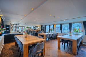Une salle à manger spacieuse et moderne, avec de grandes tables en bois et des chaises rembourrées grises, un sol imitation bois, un bar au fond, de grandes fenêtres et des compositions florales au centre de chaque table. - Ekilib