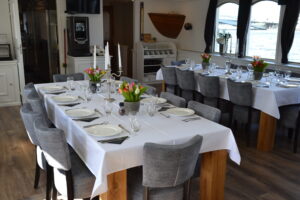 Une salle à manger où les tables sont dressées pour le repas, avec des nappes blanches, des chaises grises, des assiettes, des verres, des couverts, des compositions florales au centre des tables et de grandes fenêtres donnant sur l'eau. - Ekilib