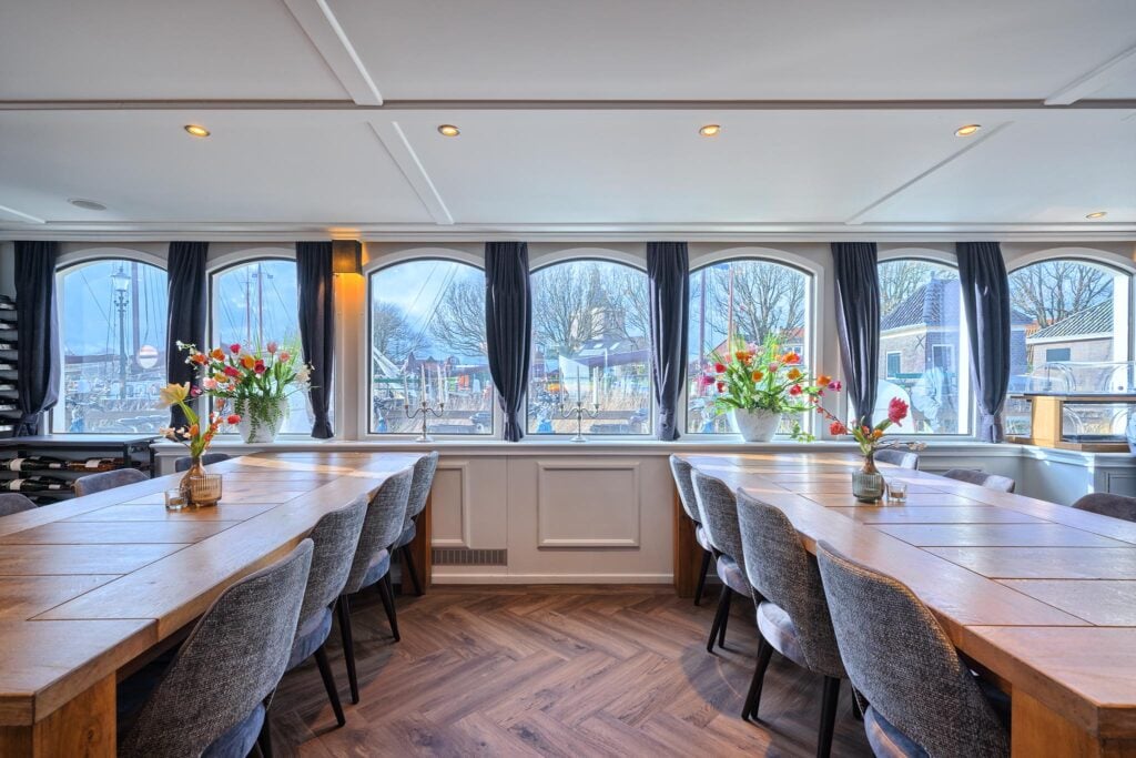 Une salle à manger avec deux longues tables en bois, des chaises rembourrées grises, des vases remplis de fleurs et de grandes fenêtres ornées de rideaux sombres, d'où l'on aperçoit le port et les bâtiments à l'extérieur. - Ekilib