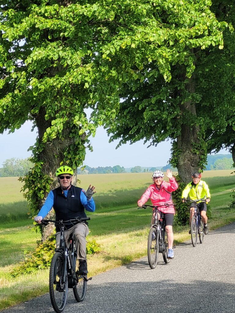 Trois personnes font du vélo le long d'une route bordée d'arbres par une journée ensoleillée. La personne de devant et celle du milieu font un signe de la main ; toutes les trois portent un casque et une veste de couleur vive. Un champ verdoyant s'étend à l'arrière-plan. - Ekilib