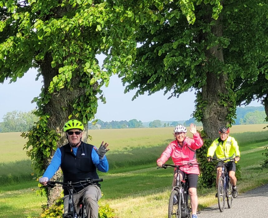 Trois personnes font du vélo le long d'une route bordée d'arbres par une journée ensoleillée. La personne de devant et celle du milieu font un signe de la main ; toutes les trois portent un casque et une veste de couleur vive. Un champ verdoyant s'étend à l'arrière-plan. - Ekilib