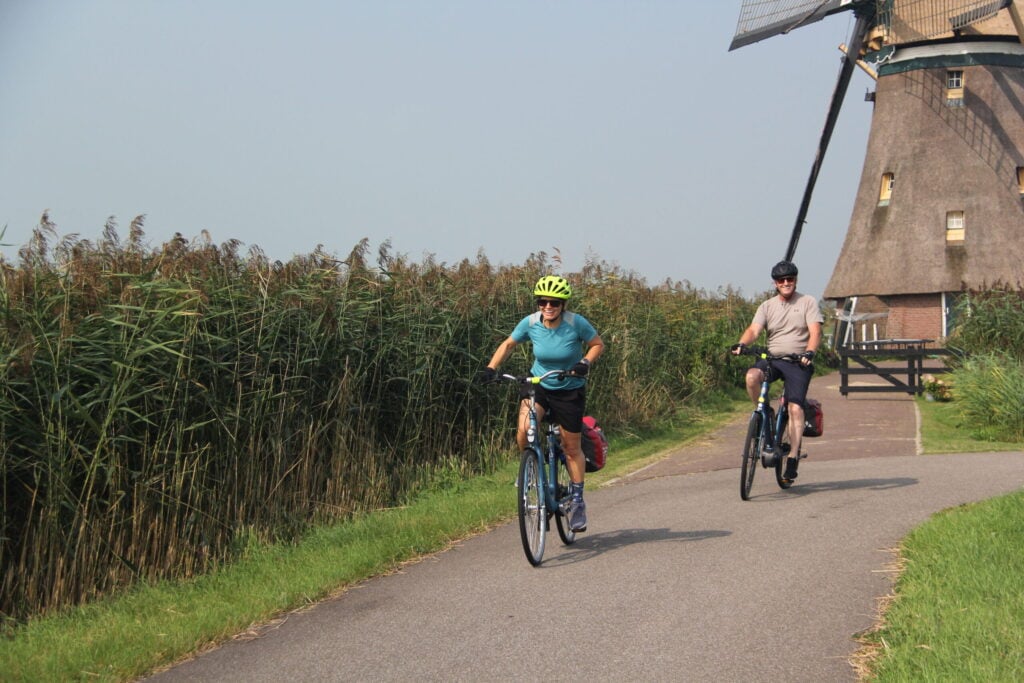 Deux personnes font du vélo sur une piste cyclable pavée, longeant des hautes herbes, avec un moulin à vent en arrière-plan sous un ciel dégagé. - Ekilib