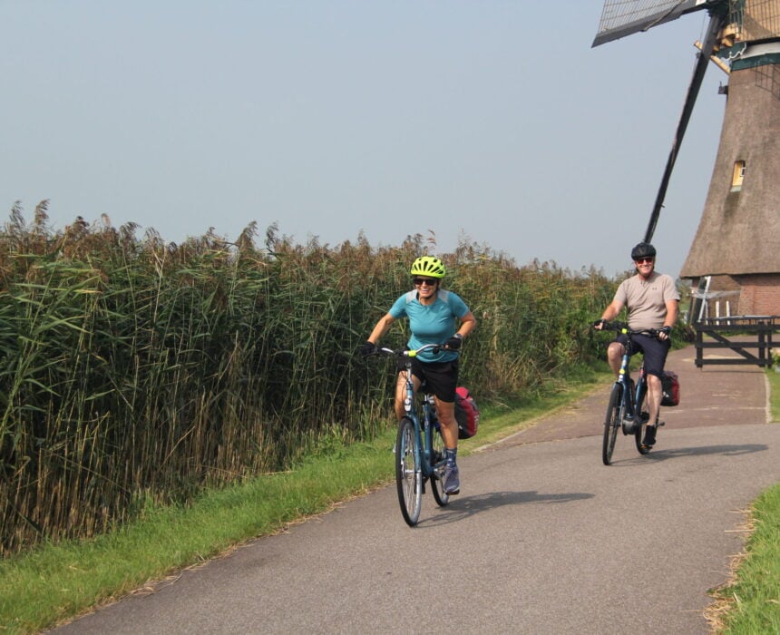 Deux personnes font du vélo sur une piste cyclable pavée, longeant des hautes herbes, avec un moulin à vent en arrière-plan sous un ciel dégagé. - Ekilib