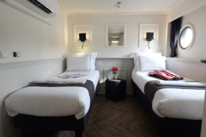 Une petite chambre avec deux lits simples, chacun recouvert de draps blancs. Une table de chevet noire, sur laquelle sont posées des fleurs et un livre, se trouve entre les deux lits. Il y a une fenêtre en forme de hublot, des appliques murales au-dessus de chaque lit et une valise sur le lit de droite. - Ekilib