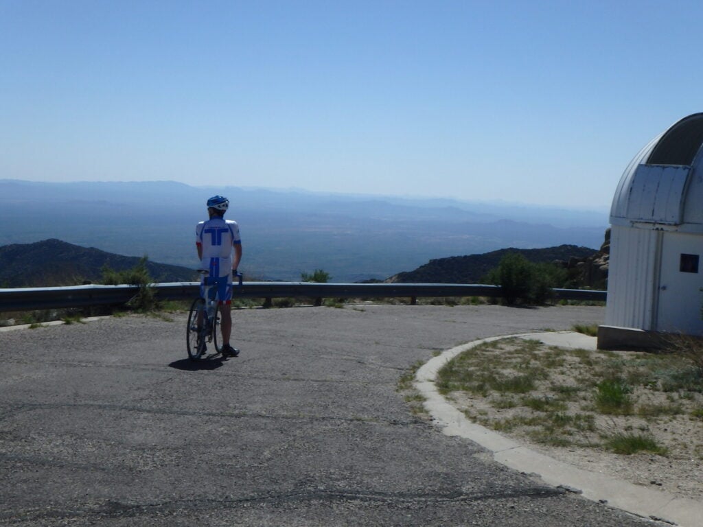 Une personne portant une tenue de cycliste se tient debout avec un vélo sur une route pavée, près d'une glissière de sécurité en métal, et regarde un paysage lointain. Un bâtiment blanc au toit arrondi se trouve à droite de l'image. - Ekilib