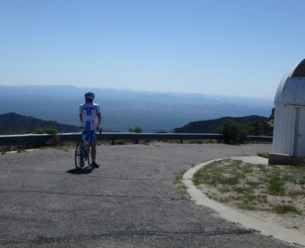 Une personne portant une tenue de cycliste se tient debout avec un vélo sur une route pavée, près d'une glissière de sécurité en métal, et regarde un paysage lointain. Un bâtiment blanc au toit arrondi se trouve à droite de l'image. - Ekilib