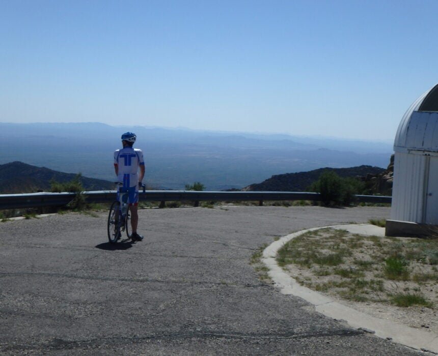 Une personne portant une tenue de cycliste se tient debout avec un vélo sur une route pavée, près d'une glissière de sécurité en métal, et regarde un paysage lointain. Un bâtiment blanc au toit arrondi se trouve à droite de l'image. - Ekilib