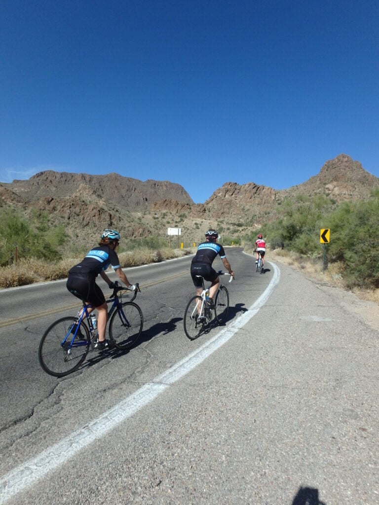 Trois cyclistes montent une route pavée avec des montagnes et une végétation désertique en arrière-plan, sous un ciel bleu clair. - Ekilib
