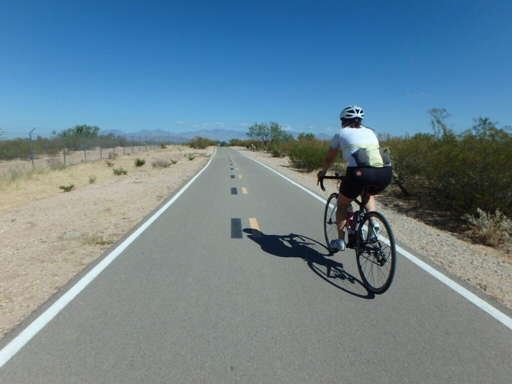 Une personne portant un casque et une tenue de cycliste roule à vélo sur une route pavée dans un paysage sec et désertique, sous un ciel bleu clair. Des arbustes et une clôture bordent la route, et des montagnes sont visibles au loin. - Ekilib