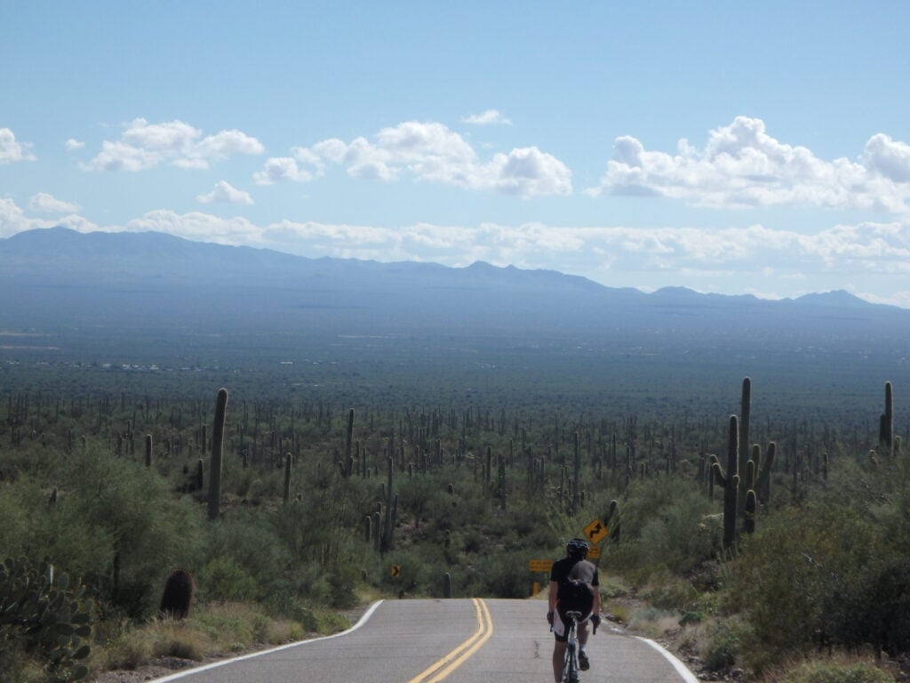 Une personne à vélo roule sur une route pavée entourée d'un paysage désertique avec des cactus, sous un ciel partiellement nuageux et une chaîne de montagnes lointaine. - Ekilib