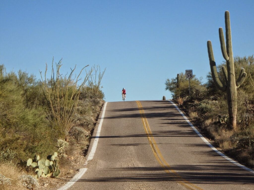 Un cycliste vêtu d'une chemise rouge monte une route pavée dans le désert, bordée de cactus et d'arbustes, sous un ciel bleu clair. Un panneau de limitation de vitesse indique 35 mph sur le côté droit de la route. - Ekilib