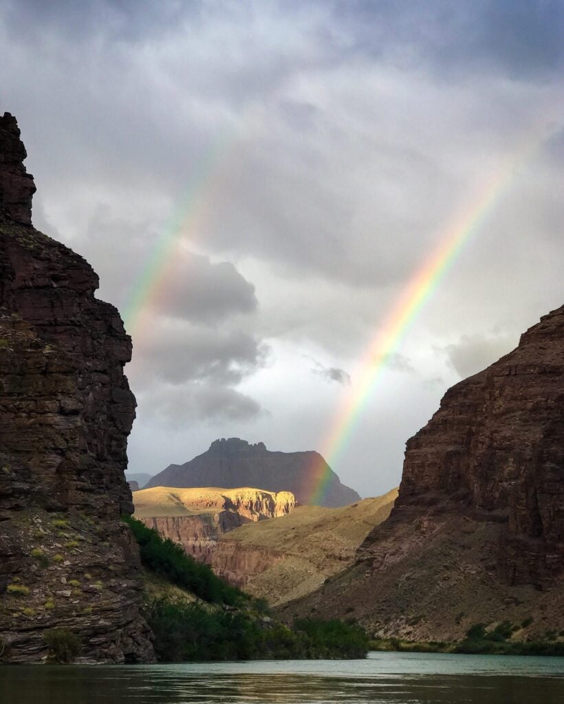 Une rivière coule entre deux grandes falaises rocheuses et un double arc-en-ciel se dessine dans un ciel nuageux. La lumière du soleil éclaire certaines parties du paysage lointain, mettant en évidence une montagne à l'arrière-plan. - Ekilib