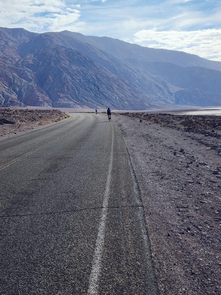 Une route pavée traverse un paysage rocheux et aride avec des montagnes en arrière-plan. Deux personnes courent ou marchent le long de la route, l'une près de la caméra et l'autre plus loin. Le ciel est partiellement nuageux. - Ekilib