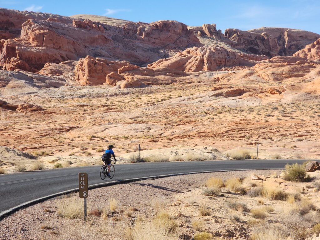 Une personne portant une veste bleue roule à vélo sur une route pavée dans un paysage désertique avec des collines rocheuses rougeâtres et une végétation clairsemée sous un ciel clair. - Ekilib
