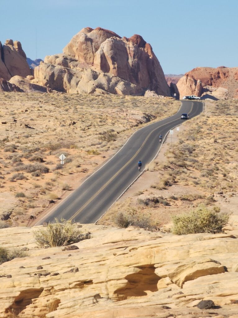 Une route goudronnée serpente en montée dans un paysage désertique avec de grandes formations rocheuses et une végétation clairsemée. Une personne marche sur la route et deux véhicules sont visibles plus loin. Le ciel est clair et bleu. - Ekilib