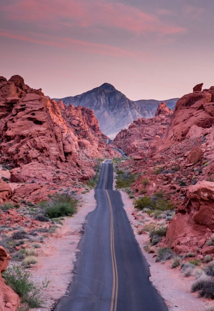 Une route pavée avec une ligne centrale jaune passe entre des formations rocheuses rouges, menant vers une montagne lointaine sous un ciel rose et violet au coucher du soleil. Une végétation désertique clairsemée borde la route. - Ekilib