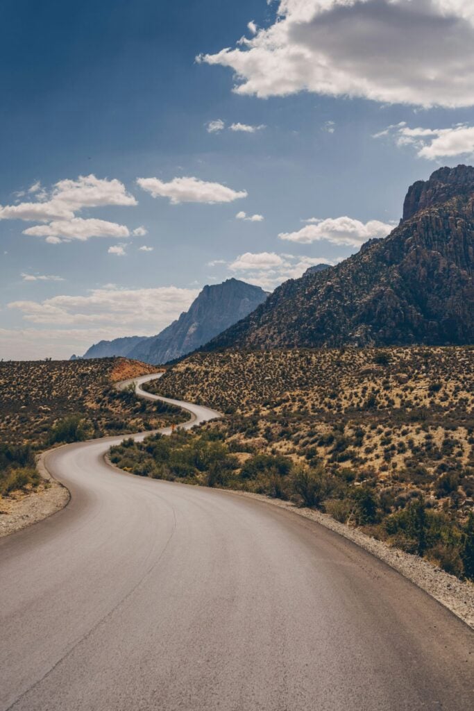 Une route pavée serpente dans un paysage sec et désertique, avec des buissons épars, des montagnes en arrière-plan et un ciel partiellement nuageux au-dessus. - Ekilib