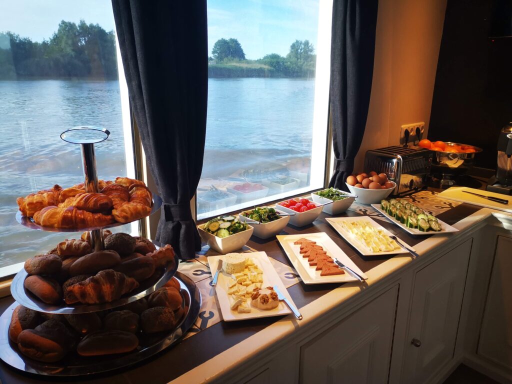 Un buffet de petit-déjeuner composé de croissants, de petits pains, de fromages, de charcuterie, d'œufs durs, de légumes, d'un grille-pain et d'un distributeur de jus de fruits est installé sur un comptoir près de grandes fenêtres donnant sur une rivière. - Ekilib