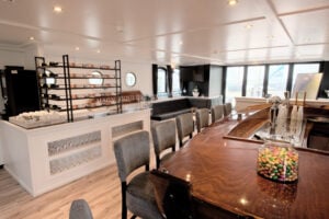 Un espace salon moderne comprend un bar en bois avec des chaises grises, un casier à vin, des canapés, une télévision à écran plat et un bocal rempli de bonbons colorés posé sur le bar. La lumière naturelle pénètre à l'intérieur par de grandes fenêtres et des hublots. - Ekilib