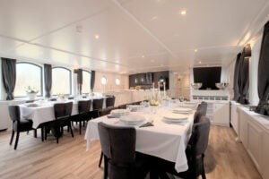 Une salle à manger dotée de grandes baies vitrées, avec des tables dressées recouvertes de nappes blanches, des chaises rembourrées noires et une décoration moderne. L'espace est bien éclairé grâce à des spots encastrés et dispose d'un parquet. - Ekilib
