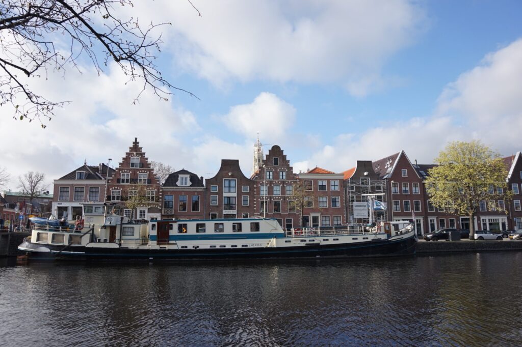 Une longue barque est amarrée sur un canal, face à des bâtiments traditionnels de style hollandais aux toits à pignon, sous un ciel partiellement nuageux. On aperçoit quelques arbres au feuillage printanier le long des berges. - Ekilib