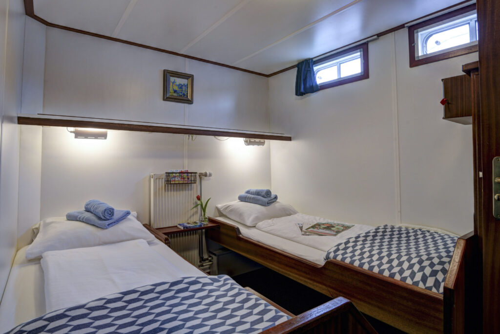 Une petite chambre avec deux lits simples, des couvertures à motifs bleus et blancs, des serviettes enroulées, un radiateur, deux petites fenêtres situées haut sur le mur et un tableau encadré au-dessus des lits. - Ekilib