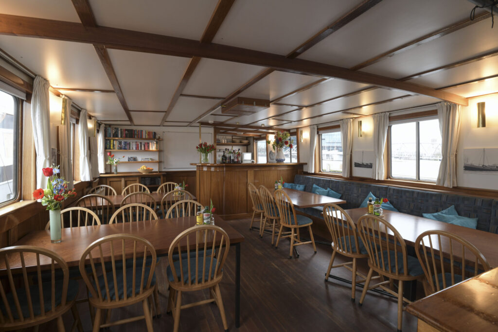 L'intérieur d'un bateau en bois comprenant des tables à manger, des chaises de style Windsor et un coin bar. Une bibliothèque est adossée au mur du fond, et de grandes baies vitrées donnent sur l'eau. Le plafond est orné de poutres apparentes et d'un éclairage encastré. - Ekilib