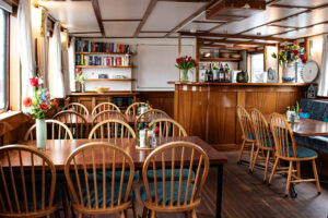 Une salle à manger chaleureuse, aux murs lambrissés, avec des tables et des chaises en bois, des vases remplis de fleurs, un bar, des étagères et des fenêtres laissant entrer la lumière naturelle. Des bouteilles et des condiments sont disposés sur les tables, et l'espace semble soigné et accueillant. - Ekilib