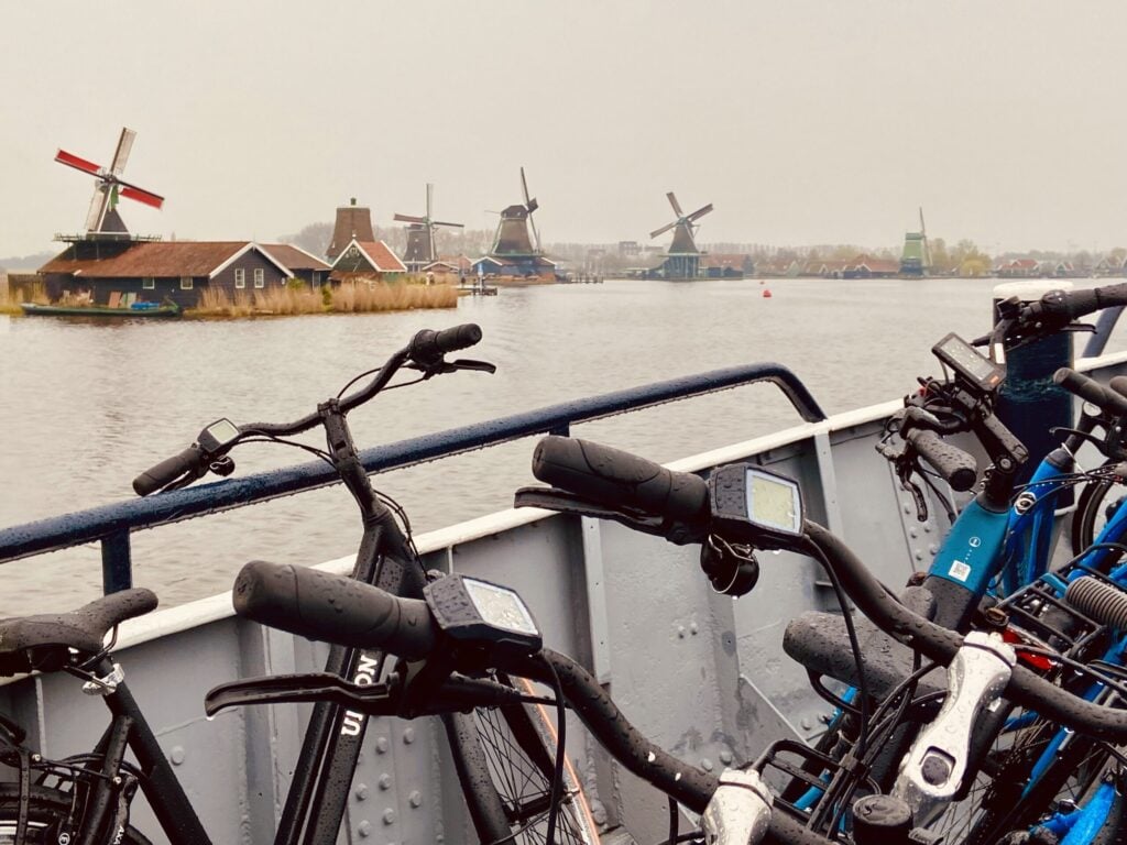 Plusieurs vélos sont garés sur un ferry, avec une rivière en arrière-plan. De l'autre côté de l'eau, on aperçoit des moulins à vent traditionnels néerlandais et des maisons par une journée nuageuse. - Ekilib