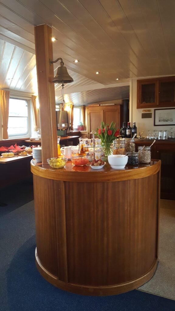 Un comptoir rond en bois accueille des bols remplis d'en-cas, des récipients en verre, des boissons et un vase de tulipes rouges, dans une pièce baignée d'une lumière chaleureuse, dotée de tables et de fenêtres, qui rappelle le salon ou la salle à manger d'un bateau ou d'un train. - Ekilib
