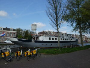 Un grand bateau blanc et gris est amarré sur un canal. Plusieurs vélos jaunes sont garés à proximité. On aperçoit à l'arrière-plan des immeubles d'habitation et un château d'eau blanc. Des arbres bordent la rive du canal. - Ekilib