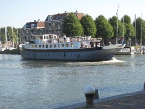 Un grand bateau blanc et gris, baptisé Mele, navigue sur un canal, longeant des voiliers amarrés et des bâtiments historiques en briques, avec des arbres qui bordent les berges. Le temps semble clair et ensoleillé. - Ekilib