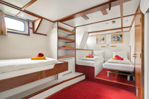 Une chambre soignée, équipée de trois lits simples, de draps blancs, de serviettes rouges et jaunes, d'étagères encastrées, d'une moquette rouge, d'une petite fenêtre et de deux photos encadrées accrochées au mur au-dessus des lits. - Ekilib