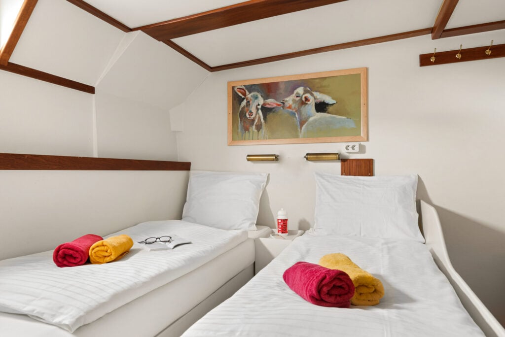 Une petite chambre avec deux lits simples, chacun recouvert de draps blancs, sur lesquels est posée une serviette enroulée, jaune et rouge. Un tableau représentant des vaches est accroché au-dessus des lits. Une paire de verres et une bouteille se trouvent sur l'un des lits. - Ekilib