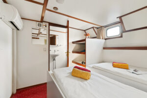 Petite chambre de type cabine équipée de deux lits simples, sur chacun desquels est posée une serviette enroulée. On y aperçoit des étagères encastrées, un miroir mural et un petit lavabo. La fenêtre et les poutres apparentes apportent de la lumière naturelle et ajoutent une touche boisée à la pièce. - Ekilib