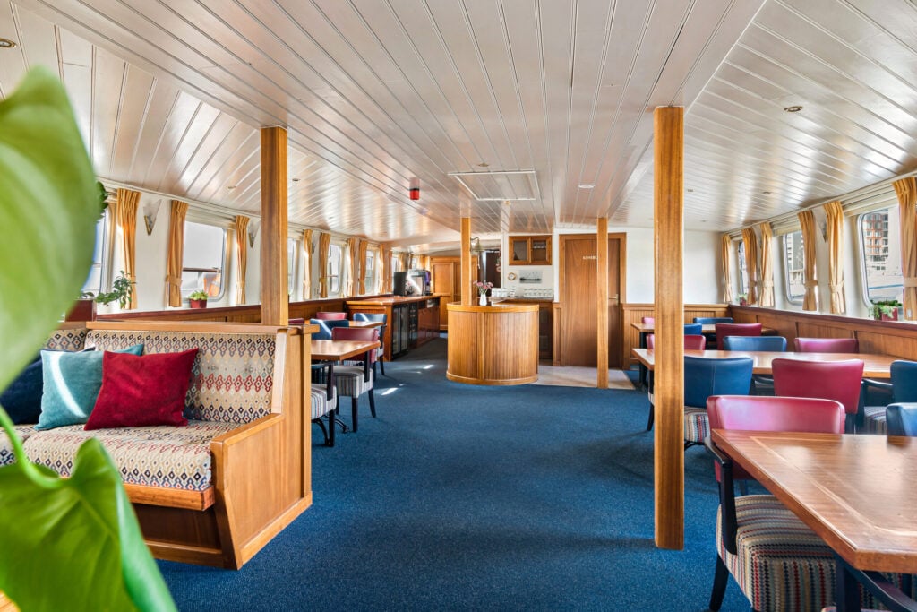 Intérieur spacieux du bateau, avec des éléments en bois, des tables, des chaises, des banquettes rembourrées, de la moquette et de grandes fenêtres ornées de rideaux jaunes. On aperçoit partiellement une plante en pot sur la gauche. La pièce est baignée de lumière naturelle. - Ekilib
