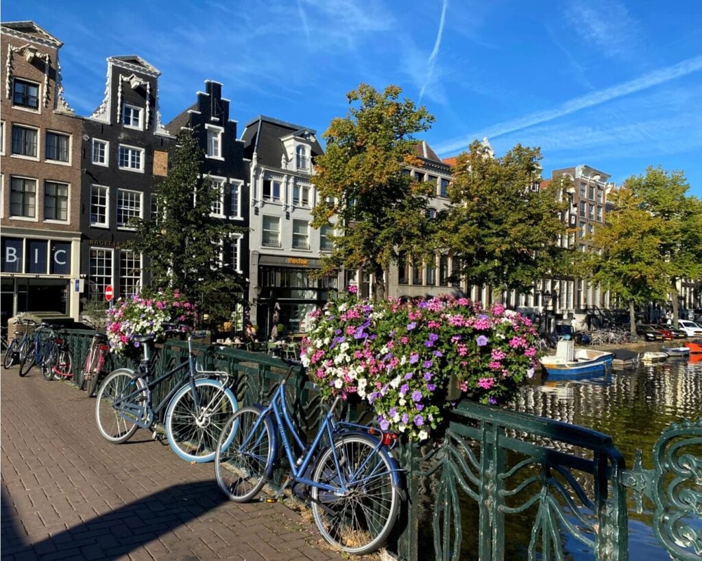 Plusieurs vélos sont garés le long d'un pont orné de jardinières, qui enjambe un canal, avec en arrière-plan des bâtiments historiques et des arbres, sous un ciel bleu limpide. - Ekilib