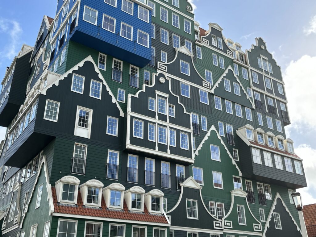 Un immeuble de plusieurs étages aux façades vertes et bleues se compose de maisons à pignons superposées de style hollandais, dotées de fenêtres aux encadrements blancs. L'architecture allie des éléments traditionnels et modernes sur fond de ciel partiellement nuageux. - Ekilib