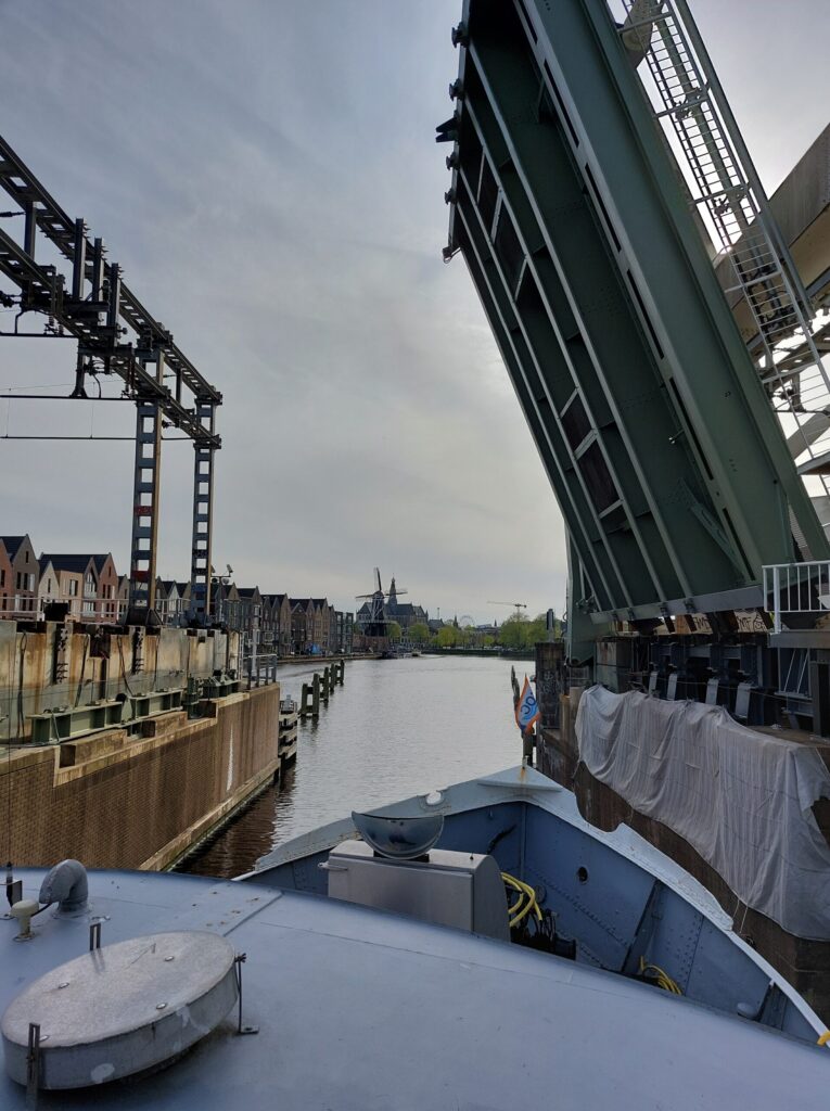 Vue depuis un bateau s'approchant d'un pont-levis surélevé enjambant une rivière, avec des bâtiments et des poteaux électriques sur la rive gauche et un moulin à vent visible au loin sous un ciel nuageux. - Ekilib