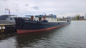 Un long bateau noir et marron baptisé « Magnifique I » est amarré dans un port. En arrière-plan, sous un ciel nuageux, on aperçoit des bâtiments modernes et des grues industrielles. - Ekilib