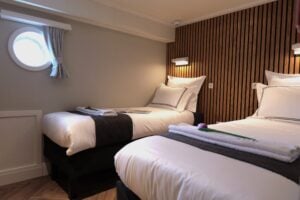 Deux lits simples soigneusement faits, avec des draps blancs et des chemins de lit foncés, dans une chambre dotée d'un mur d'accent lambrissé et d'une petite fenêtre ronde. Sur chaque lit sont posés une serviette et une fleur. - Ekilib