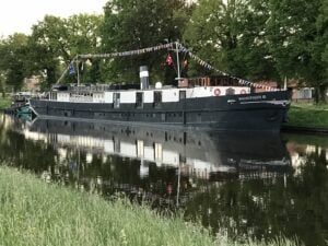 Un grand bateau noir et blanc, baptisé « Magnifique II », est amarré le long d'un canal paisible, orné de guirlandes de drapeaux colorés. Des arbres verts et de l'herbe bordent la rive, et le bateau se reflète dans l'eau. - Ekilib