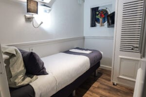 Un petit lit soigneusement fait, avec une serviette pliée et un oreiller, dans une chambre compacte aux murs blancs, dotée d'un parquet, d'une applique murale et d'une fenêtre ronde d'où l'on aperçoit un pot de fleurs à l'extérieur. - Ekilib