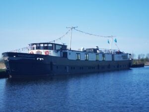 Un grand bateau fluvial noir et blanc, baptisé « Magnifique IV », est amarré sur des eaux calmes, sous un ciel bleu limpide. Le bateau est orné de pavillons de signalisation et deux drapeaux bleus flottent sur son pont. On aperçoit des arbres à l'arrière-plan. - Ekilib