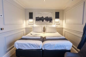 Une chambre d'hôtel avec deux lits jumeaux soigneusement faits et rapprochés, du linge de lit blanc, deux serviettes pliées, une photo d'une ville au-dessus de la tête de lit et des appliques murales de chaque côté. - Ekilib